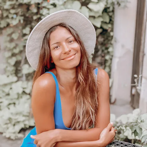 Smiling woman with long hair wearing a white hat and blue sleeveless top, sitting outdoors near green plants.